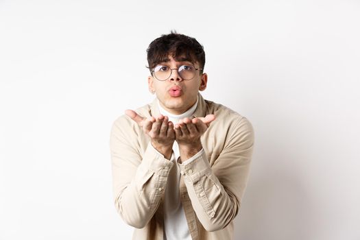 Cute boyfriend sending air kiss at camera on happy Valentines day, looking tender at lover, standing in casual clothes and glasses on white background.