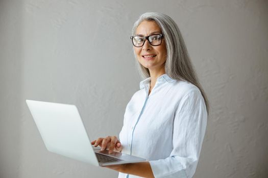 Positive hoary haired middle aged Asian woman in white blouse touches pad of laptop standing near grey stone wall in studio