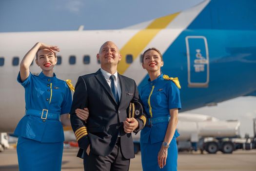 Happy pilot and two pretty stewardesses standing together in front of an airplane and smiling away after landing. Aircraft, aircrew, occupation concept