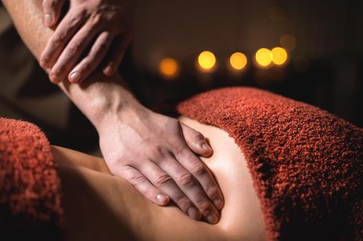 Professional premium massage in a dark atmospheric cabinet. Close-up hands Young man doing massage to a female client in a dark office on the background of burning candles.