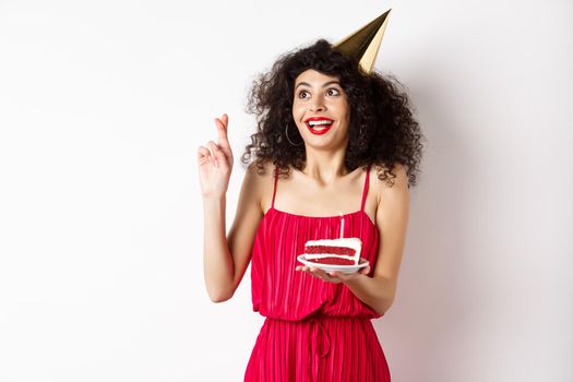 Excited birthday girl making wish, cross fingers good luck and looking aside at logo, celebrating bday, holding piece of cake, white background.