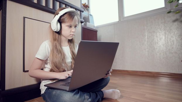 A smart little girl of seven years old in white headphones with a laptop in her hands is pushing on the floor in her room. The young generation on the Internet and IT technology.
