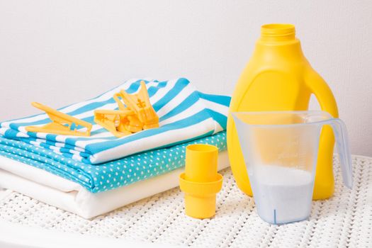 a stack of colored laundry, clothespins, laundry detergent in a measuring cup and washing gel in a yellow bottle on a white plastic basket for dirty laundry, washing colored clothes