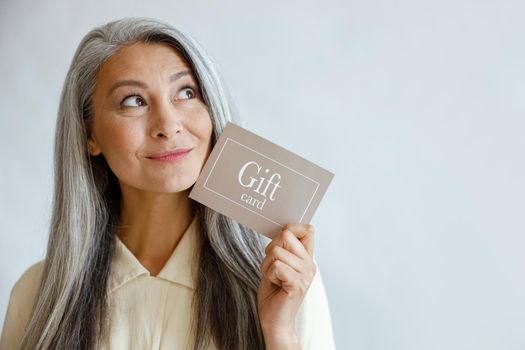 Dreamful grey haired Asian woman holds gift card near face standing on light grey background in studio, space for text. Shopping certificate