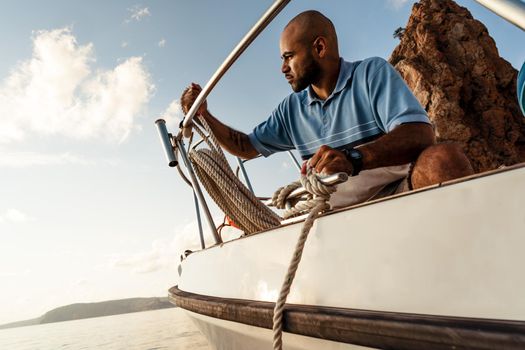 Young african american sailor tying ropes on sailboat in the sea on sunset, close up