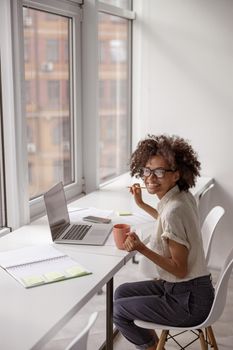 Happy Afro American lady sitting near the window while using laptop and enjoying cup of drink