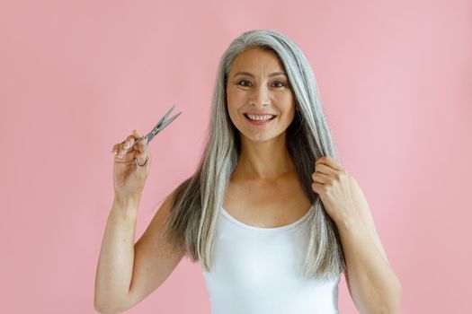 Cheerful middle aged Asian lady holds lock of long silver hair and scissors on pink background in studio. Mature beauty lifestyle