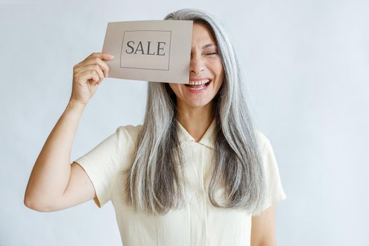 Laughing middle aged woman with long hoary hair covers eye with Sale sign posing on light grey background in studio