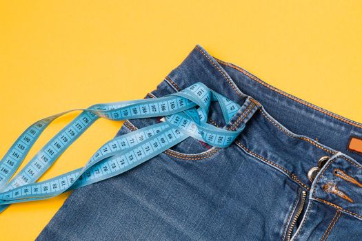 blue measuring tape is threaded into jeans instead of a belt, blue jeans on a yellow background, copy space, weight loss and weight control concept, measuring body volume