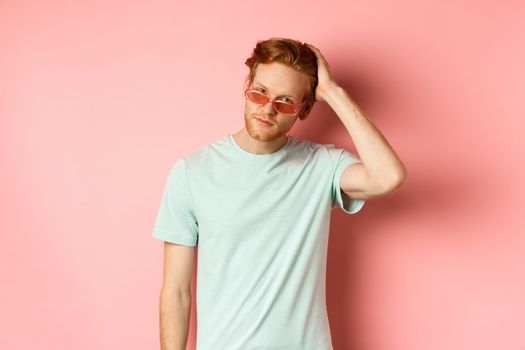 Handsome young redhead man in sunglasses, brushing hair with hand and looking smug and confident at camera, standing over pink background.