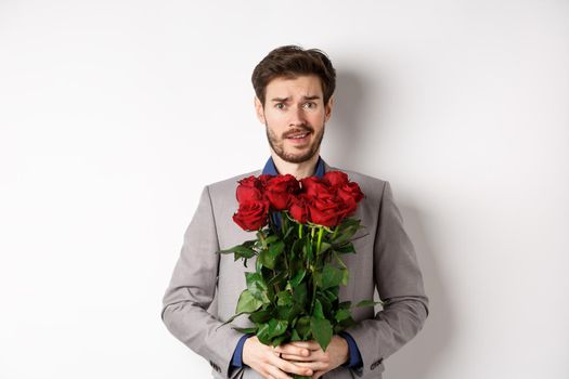 Worried boyfriend in suit, holding flowers roses and looking doubtful at camera, standing with bouquet on valentines day against white background.