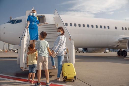 Back view of mother with two little kids wearing protective mask boarding the plane in the daytime, ready for summer vacations. People, traveling, vacation concept