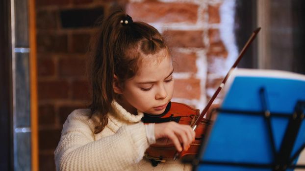 A little concentrated girl in white sweater playing violin by notes. Mid shot