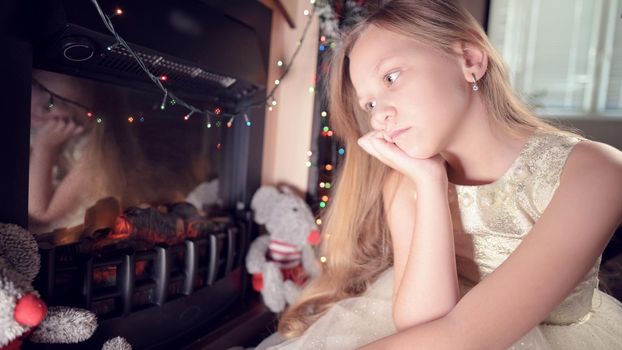 A little sad disappointed girl sits by an artificial fireplace and is sad next to soft toys. The concept of spoiled Christmas holiday.