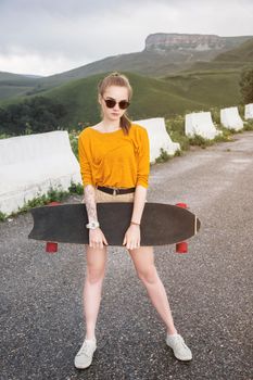 Beautiful and fashionable young woman in sunglasses and with a tattoo poses with a skateboard or longboard against the sunset sky.