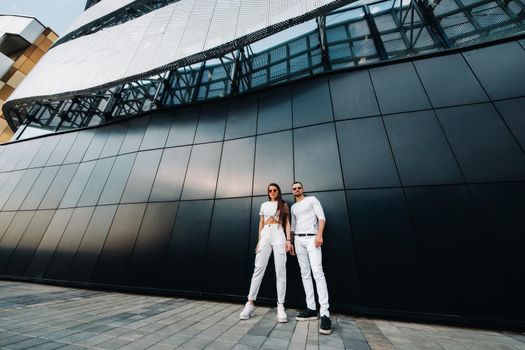 A girl and a guy walk around the city in white clothes and glasses, a couple in love against a black wall
