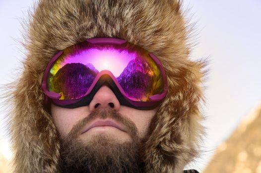 Close-up portrait of a bearded happy snowboarder skier in a ski mask with goggles and a fur big old-school hat. on a background of a winter snowy mountains.