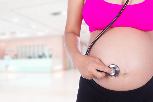 close-up of pregnant woman with stethoscope listening belly to baby in hospital background