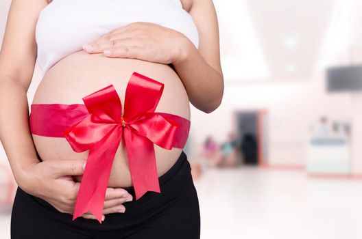 close up pregnant woman with red ribbon gift on belly in hospital background