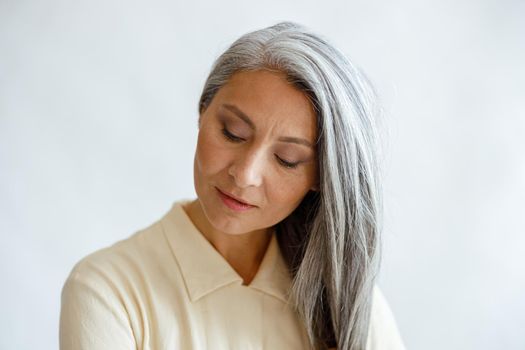 Tranquil middle aged Asian woman in elegant blouse with loose silver hair posing on light background in studio. Mature beauty lifestyle
