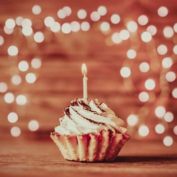 Holiday cream cupcake with burning candle on a wooden table against blurred lights.