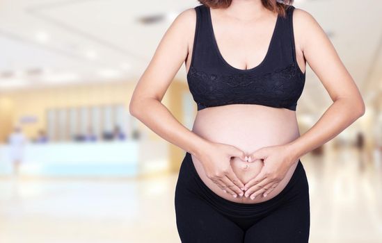 Pregnant Woman holding her hands in a heart shape on her belly in hospital background