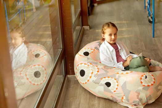 Full body of adorable girl in casual wear looking away while sitting in comfortable bean bag near window in building