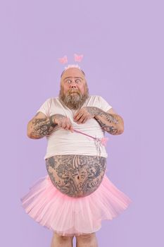 Shocked bearded man with overweight wearing funny fairy costume holds magic stick standing on purple background in studio
