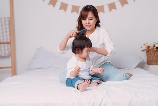 Mother combing her little daughters hair on the bed in bed room.