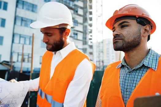 Two young male engineers in uniform and hardhats working at construction site, close up