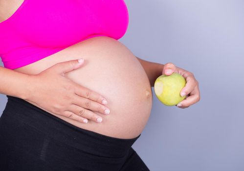 close-up belly of pregnant woman with green apple on gray wall background