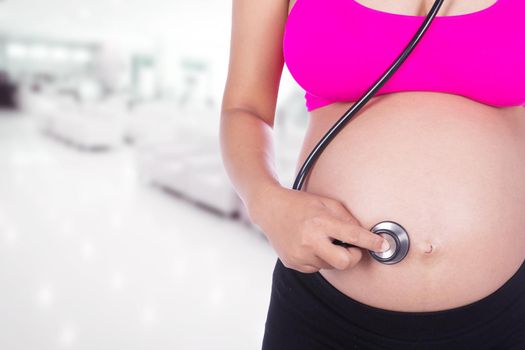 close-up of pregnant woman with stethoscope listening belly to baby in hospital background