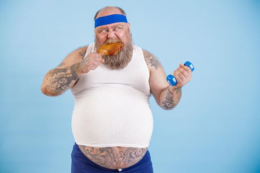 Funny plus size man eats chicken leg and does sports exercises with dumbbell standing on light blue background in studio