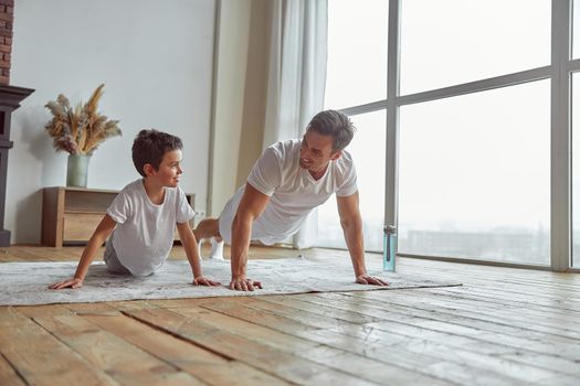 Low angle of jolly son enjoying workout with dad and doing plank together near window