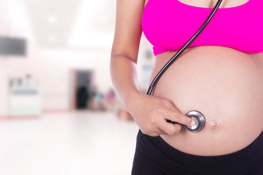 close-up of pregnant woman with stethoscope listening belly to baby in hospital background