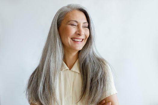 Pretty laughing Asian lady with natural silver hair poses for camera on light grey background in studio. Mature beauty lifestyle