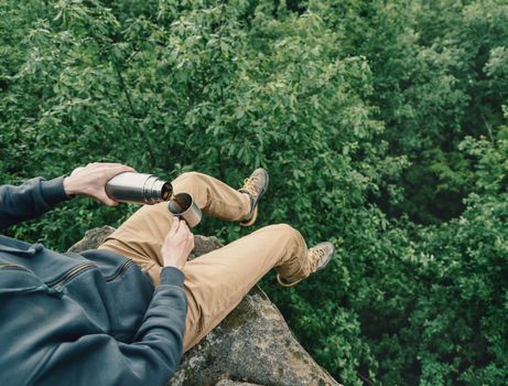 Traveler young man sitting on top of cliff over forest and pouring tea from thermos to cup. Point of view shot