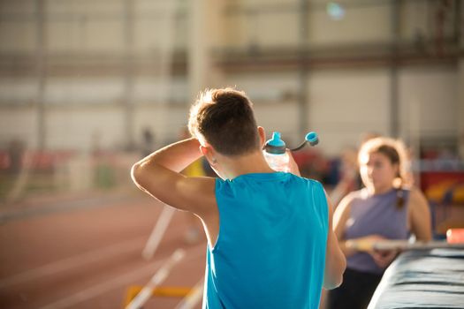 Young man in blue shirt standing on the sports stadium holding a water bottle - wiping out the sweat from his forehead. Mid shot