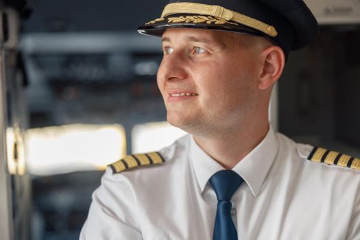 Close up portrait of cheerful male pilot in uniform and hat smiling away while posing, standing inside of the airplane. Transportation, aircrew concept