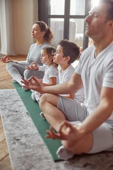 Calm parents with daughter and son are sitting on mat in lotus position and doing mental practice