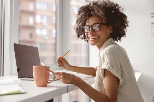 Smiling pretty Afro American lady wearing glasses and working with paper and laptop