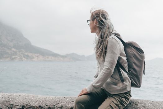 Traveler young woman sitting on coast near the sea and enjoying view of nature at windy weather