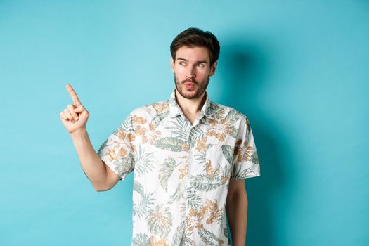 Handsome guy wearing summer shirt on vacation, pointing and looking aside at empty space, making amazed face, standing on blue background.