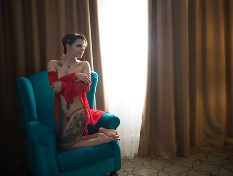 Young woman with tattoos in a sexy red underwear sitting in the chair on her knees. Mid shot