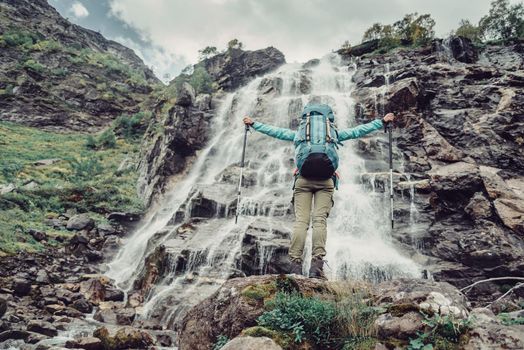 Happy hiker young woman standing with raised arms in front of waterfall