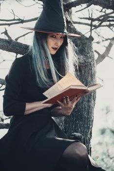 Young woman in costume of Halloween witch sitting with book on tree in autumn forest