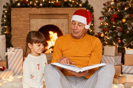 Grandfather wearing orange sweater and santa claus hat reading book to his cute granddaughter while sitting in festive living room near fireplace and Christmas tree.