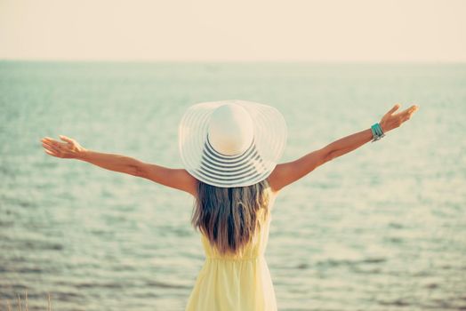 Young woman wearing in dress and hat with a wide brim standing with raised arms and enjoying view of sea, rear view. Toned image