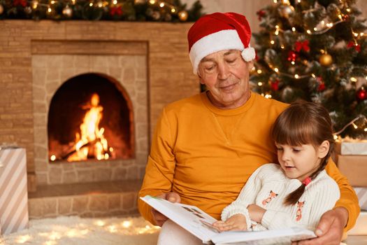 Positive senior man wearing orange sweater and santa claus hat reading fairy tale to his charming granddaughter, posing in living room with fireplace and Christmas tree.