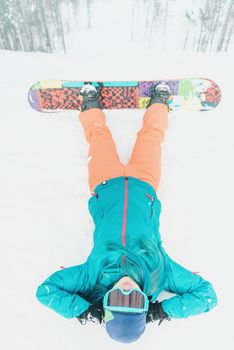 Young woman with snowboard lying on snow in front of the slope, top view